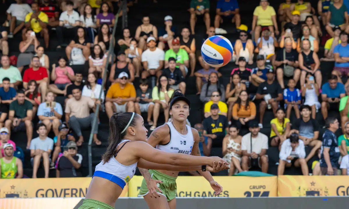 Duplas do vôlei de praia do Brasil em ação durante o Pré-Olímpico, com uma atleta cortando a bola diante da torcida